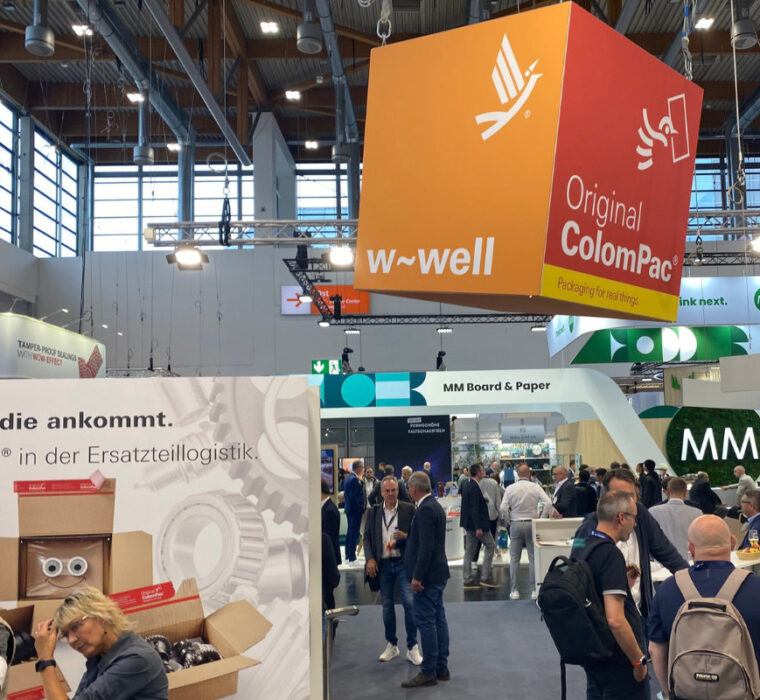 Messehalle mit ColomPac®-Stand zur Ersatzteillogistik; orangefarbener Würfel mit „w-well“ oben sichtbar.