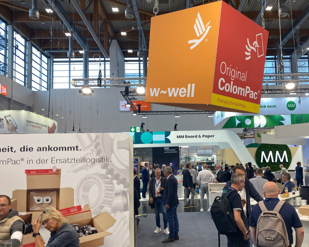 Messehalle mit ColomPac®-Stand zur Ersatzteillogistik; orangefarbener Würfel mit „w-well“ oben sichtbar.